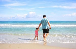 A father holds his daughter's hand while walking on the beach.